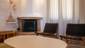 A cozy living room with a fireplace, two wicker chairs, a round table, and a large window with curtains.