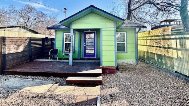 Une petite maison à un étage, avec une façade verte et une porte violette. Elle possède une terrasse en bois et une chaise à l’extérieur.