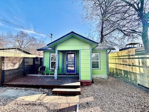 Una casa pequeña de un solo piso con fachada verde y puerta morada. Tiene una terraza de madera y una silla afuera.