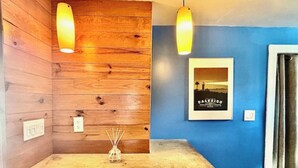 Un mur en bois orné d’un tableau encadré, une table sur laquelle se trouve un récipient en verre, et deux lampes suspendues.