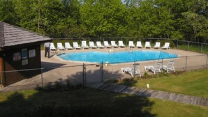 Une piscine rectangulaire entourée de chaises longues blanches, d’une terrasse en bois et d’une zone délimitée par une clôture, avec des arbres en arrière-plan.
