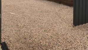 Une allée de gravier mène à un bâtiment entouré d’une clôture en métal et agrémenté de plantes.