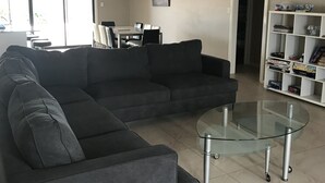 Un salon moderne avec un canapé d’angle, une table basse en verre et une bibliothèque.
