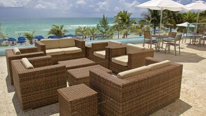 A beachside lounge area with wicker furniture and white umbrellas, overlooking a pool and the ocean.