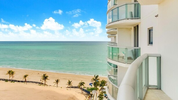 A beachfront hotel with a balcony overlooking the ocean, a sandy beach, and palm trees.