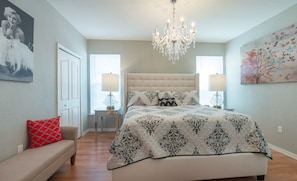 Un dormitorio con una cama grande, dos lámparas de noche, un candelabro, un banco con un cojín rojo y un cuadro de una mariposa en la pared.