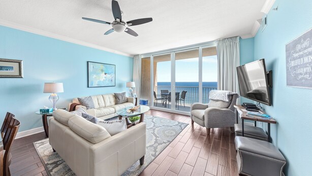 A spacious living room with a large window offering a view of the ocean, a ceiling fan, and a flat-screen TV mounted on the wall.
