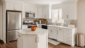 Une cuisine moderne avec des meubles blancs, des appareils en acier inoxydable et un îlot central agrémenté d’un panier de fruits.