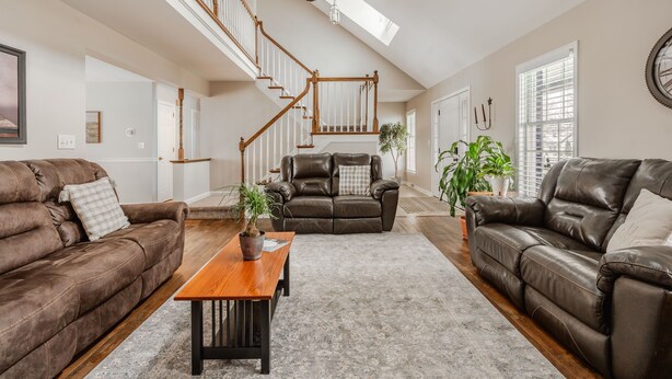 Un salon spacieux avec un plafond haut, un escalier en bois, des canapés en cuir et une grande fenêtre qui laisse entrer la lumière naturelle.