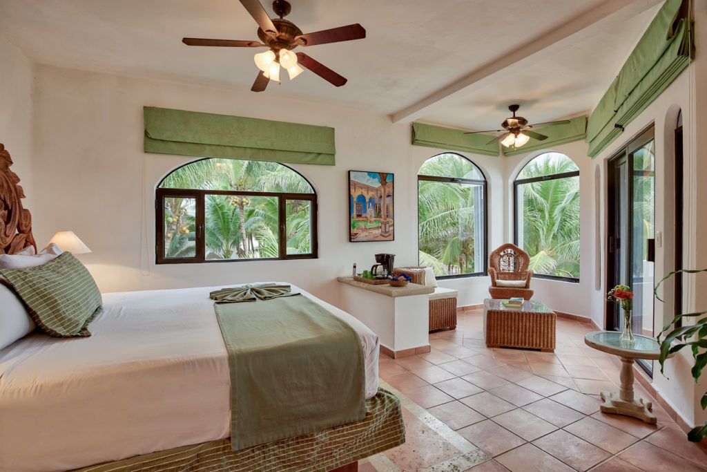 A spacious room with a large bed, two ceiling fans, a small table, and a chair. There are large windows with views of palm trees.