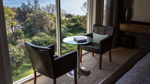 Habitación con un ventanal grande que ofrece vistas a un paisaje natural y al mar. Hay una mesa redonda de cristal con un libro y dos sillones de cuero.