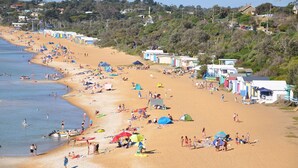 Una playa con numerosas tiendas de campaña y cabañas, una orilla arenosa, y un fondo de casas y árboles.
