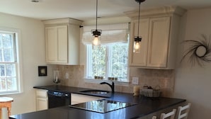 Una cocina con una isla central, muebles blancos, iluminación colgante y vistas a árboles por la ventana.