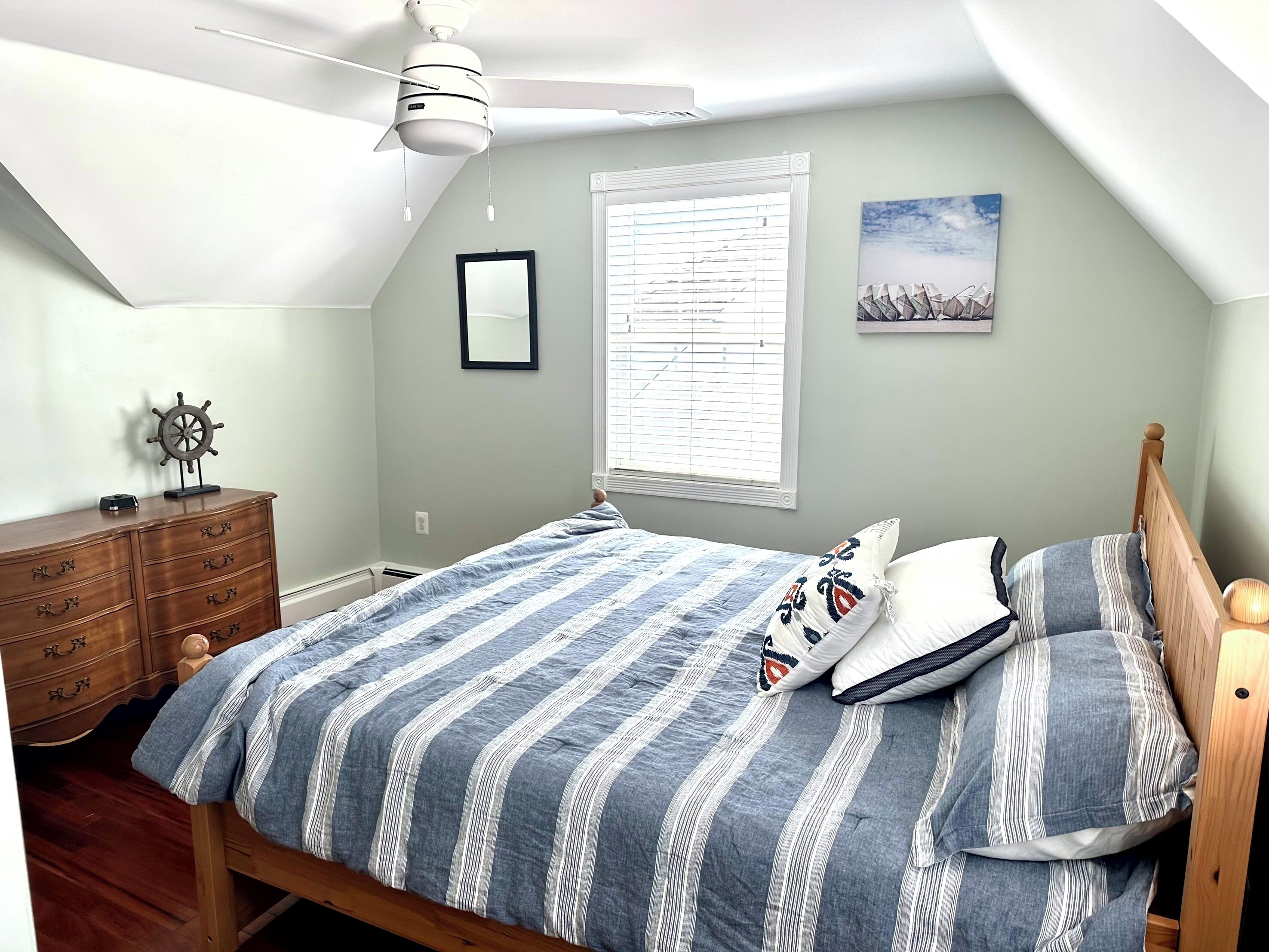 A bedroom with a sloped ceiling, a bed with striped bedding, a wooden dresser, a ceiling fan, a window with blinds, and a framed picture on the wall.