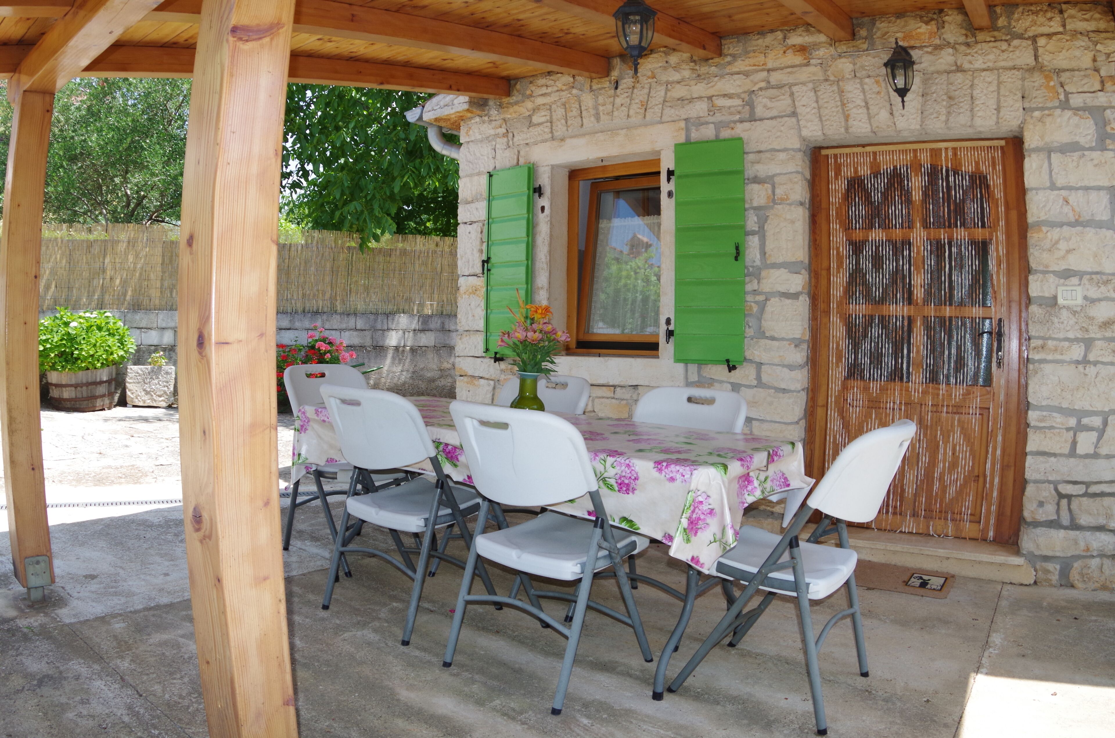 Een terras buiten met een stenen muur, houten balken, een tafel met stoelen en een houten deur met groene luiken.