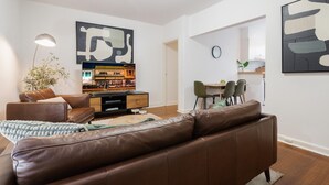 Un salon moderne avec un canapé en cuir marron, une table basse et un téléviseur posé sur un meuble en bois. On y trouve également un coin repas avec une table et des chaises, ainsi qu’un grand tableau abstrait accroché au mur.