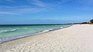 A wide sandy beach with gentle waves, a clear blue sky, and distant buildings. - Beachfront Condo - 3 BR- Heated Pool - Free Boat Docks – Upscale – Siesta Key (Siesta Key)
