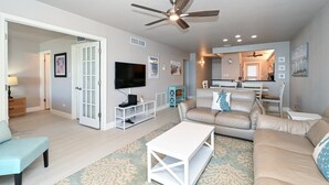 A living room with a beige sofa, a white coffee table, a TV, and a ceiling fan. - Beachfront Condo - 3 BR- Heated Pool - Free Boat Docks – Upscale – Siesta Key (Siesta Key)