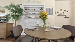 Un coin repas avec une table ronde dressée pour quatre personnes, entourée de chaises. On aperçoit en arrière-plan une bibliothèque avec des objets décoratifs et un tableau affichant le menu.