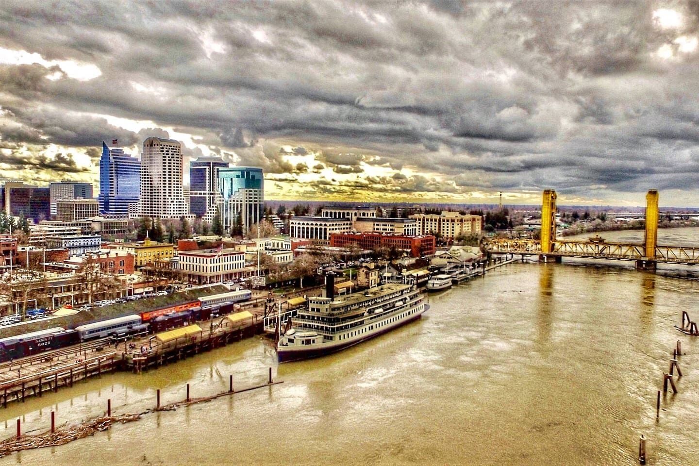 A river cityscape with a prominent bridge, a river with boats, and a dense urban area with various buildings.