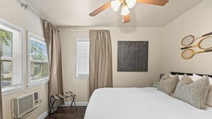 Une chambre avec un grand lit, deux fenêtres équipées de stores, un ventilateur de plafond, un climatiseur fixé au mur et un portant à vêtements.
