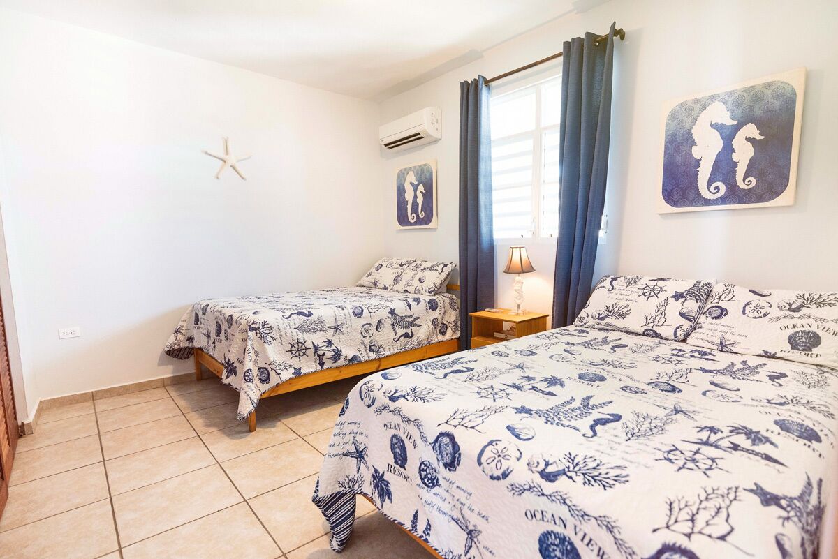 A bedroom with two beds, each with a nautical-themed bedspread, a small bedside table with a lamp, and a window with curtains.