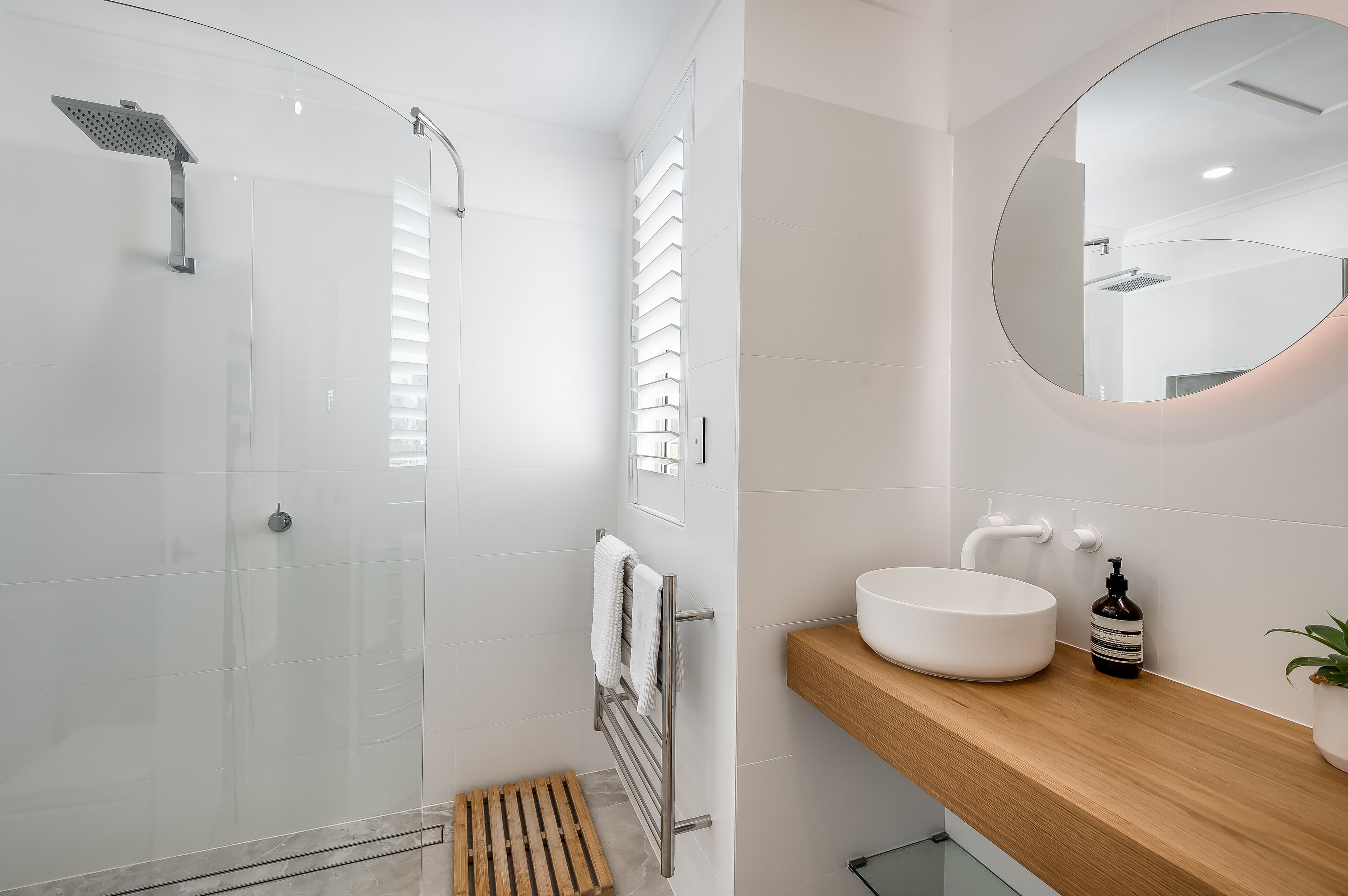 Een moderne badkamer met een douche met glazen wanden, een houten wastafelmeubel en een ronde spiegel.