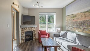 A living room with a stone fireplace, a flat-screen TV, a red armchair, a grey sofa, and a large painting on the wall. -  Location, Location, Location! Large Corner Ground Floor Newly Renovated Condo (Silver Star Mountain)