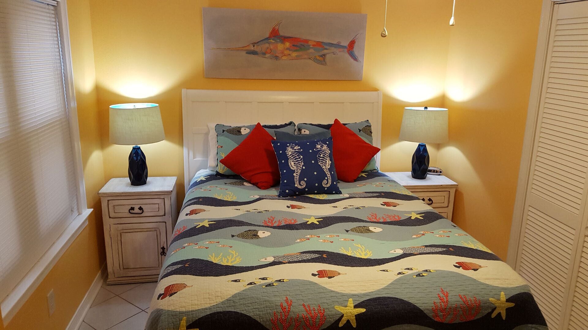 Un dormitorio con cama, dos mesitas de noche, una mesita auxiliar, una ventana con persianas y un cuadro en la pared.