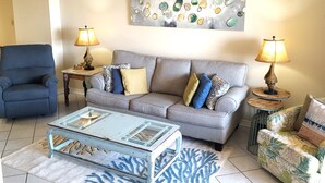 Un salon avec un canapé gris, deux tables d’appoint avec des lampes, un fauteuil bleu et une table basse en verre.