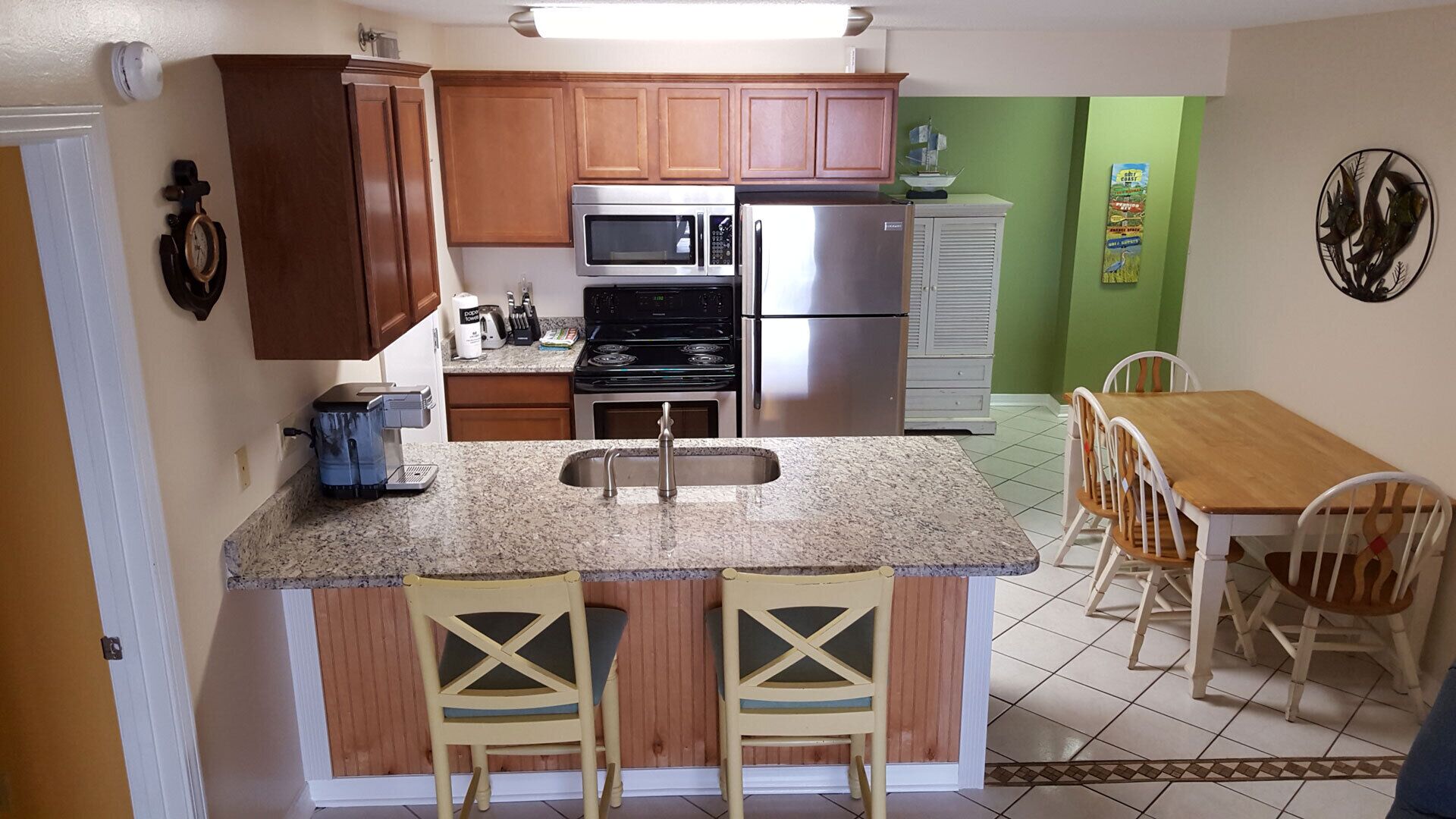 A compact kitchen with wooden cabinets, a stainless steel refrigerator, a microwave, and a coffee maker. There is a small dining area with a wooden table and chairs.