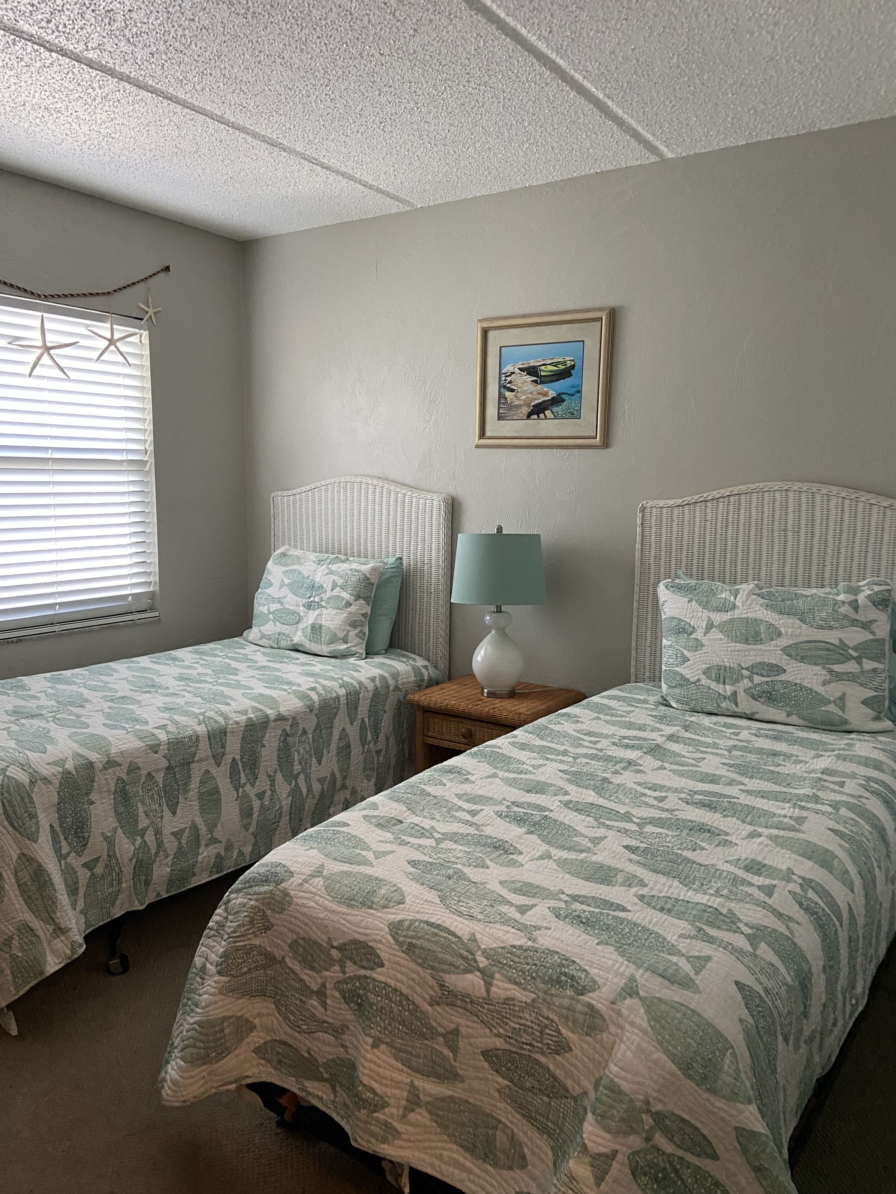 Un dormitorio con dos camas individuales, una mesita de noche con lámpara, un cuadro enmarcado en la pared y una ventana con persianas.