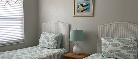 Un dormitorio con dos camas individuales, una mesita de noche con lámpara, un cuadro enmarcado en la pared y una ventana con persianas.