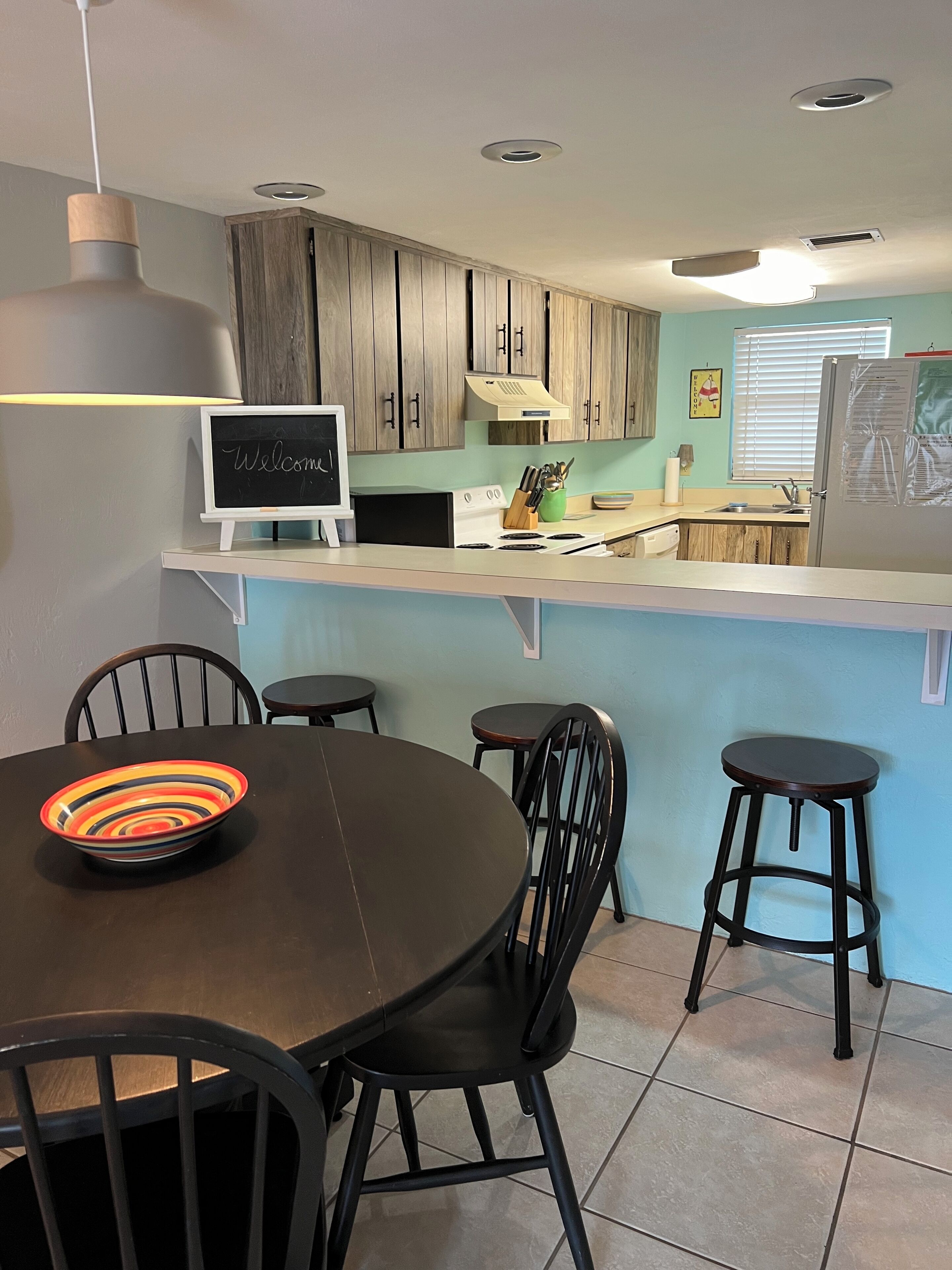 A modern kitchen with wooden cabinets, a white countertop, and a chalkboard sign. There is a dining area with a round table and four chairs.