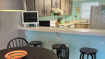 A modern kitchen with wooden cabinets, a white countertop, and a chalkboard sign. There is a dining area with a round table and four chairs.