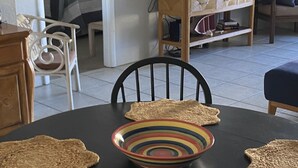 Uma área de refeições com uma mesa preta, dois jogos americanos trançados, uma tigela colorida e uma mesa lateral de madeira com livros.