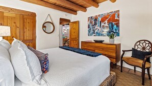 A bedroom with a large bed, wooden ceiling beams, a wooden dresser, a chair, and a painting on the wall.