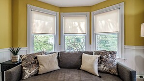 A corner room with a gray sofa, three windows with white blinds, and a small table with a plant. - Fall In Love With Portland! (Portland)