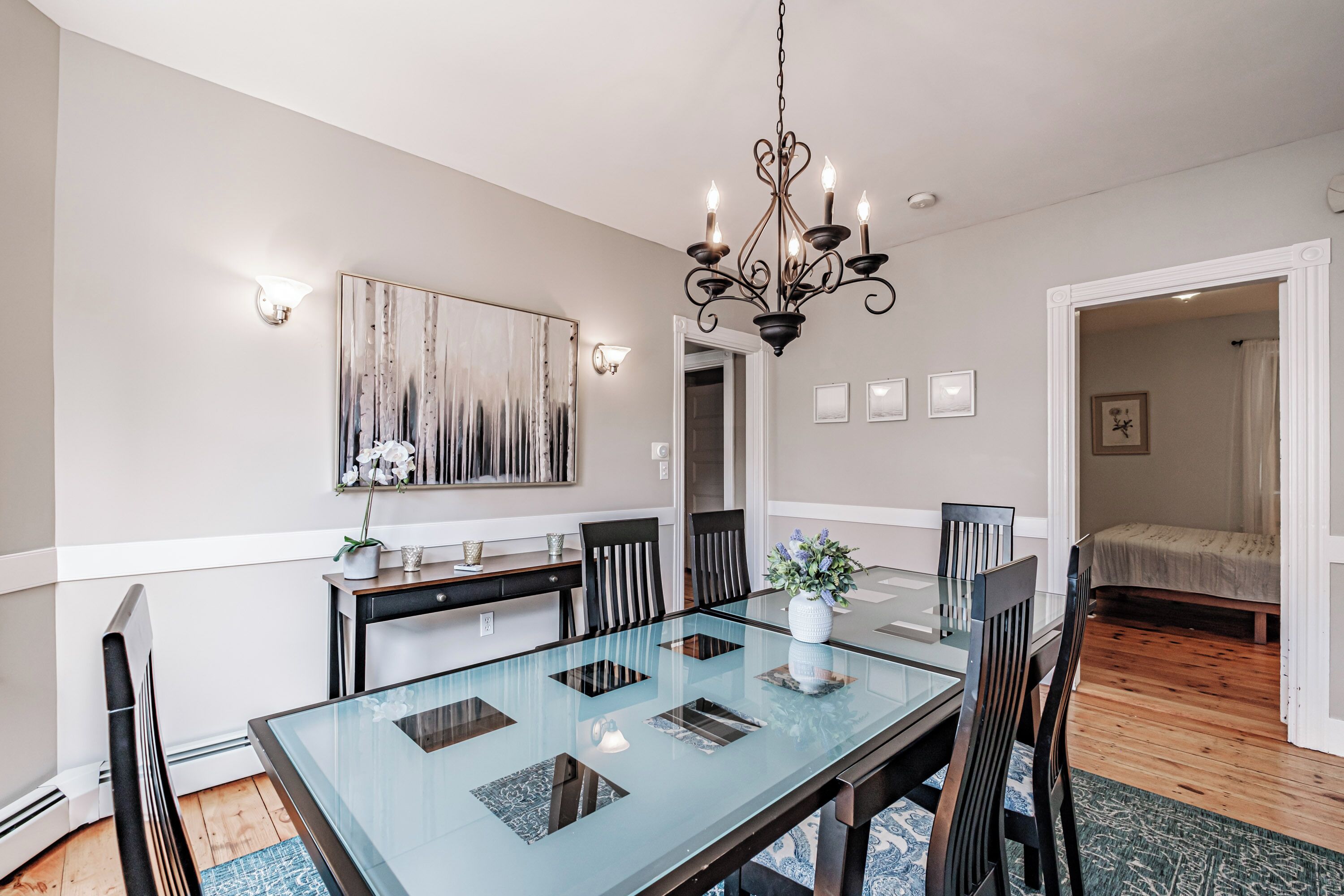 A dining area with a glass table, black chairs, a chandelier, and a painting on the wall.