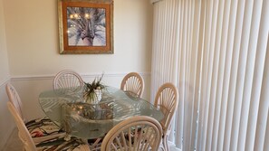 A dining area with a glass table surrounded by wicker chairs, a framed picture on the wall, and a window with vertical blinds. - Berm Bay un 22 Gorgeous Townhouse , White Sandy Beaches, elevator, heated pool (Bradenton Beach)