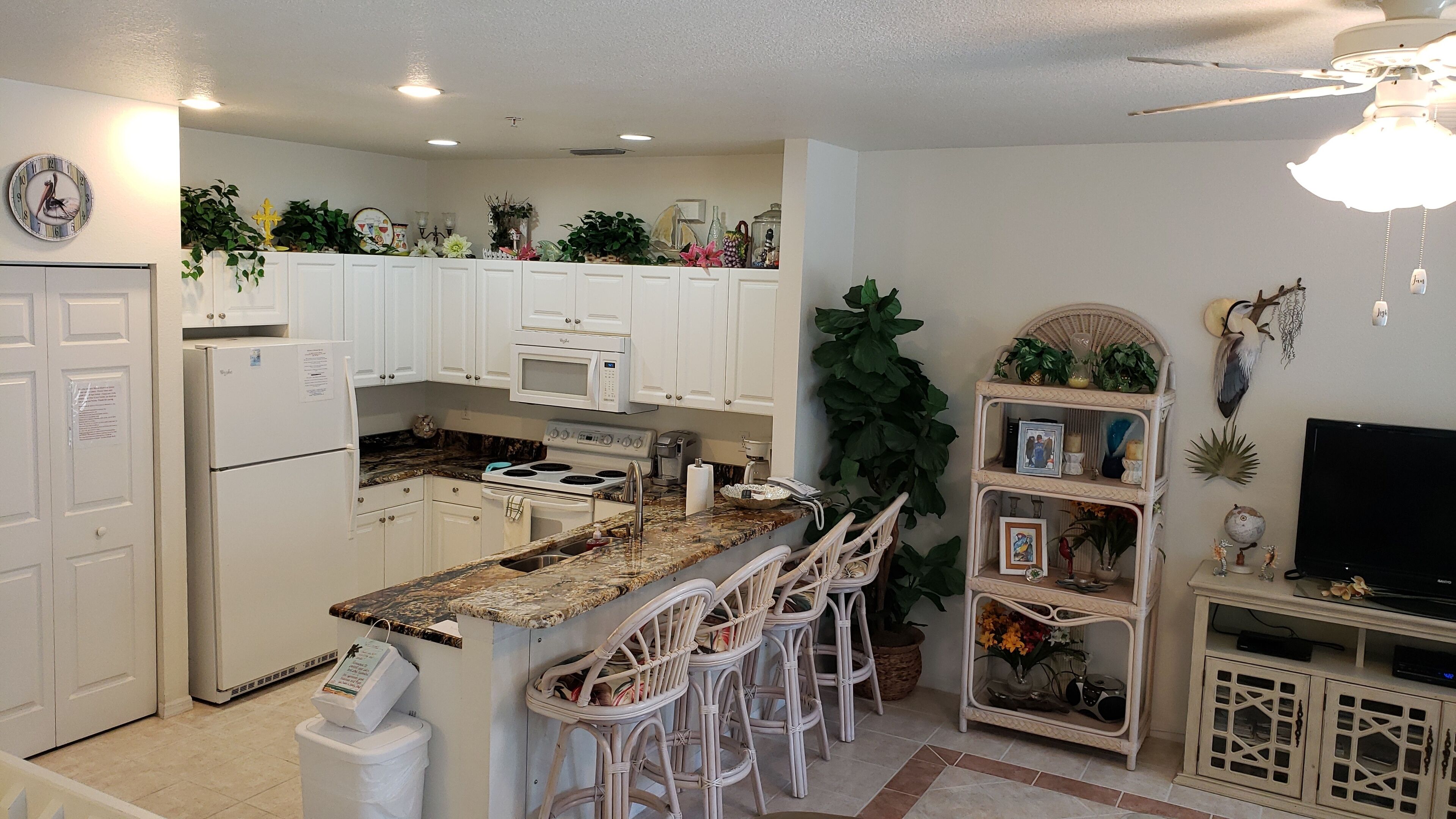 A kitchen with white cabinets, a microwave, a stove, and a sink. There are white bar stools and a small shelf with decorative items.