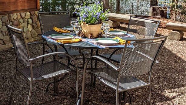 An outdoor dining area with a table set for four, surrounded by metal chairs, a wooden table, and a potted plant with blue flowers.