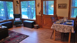 A dining area with a table set for four, wooden floors, a patterned rug, and large windows with a view of trees.