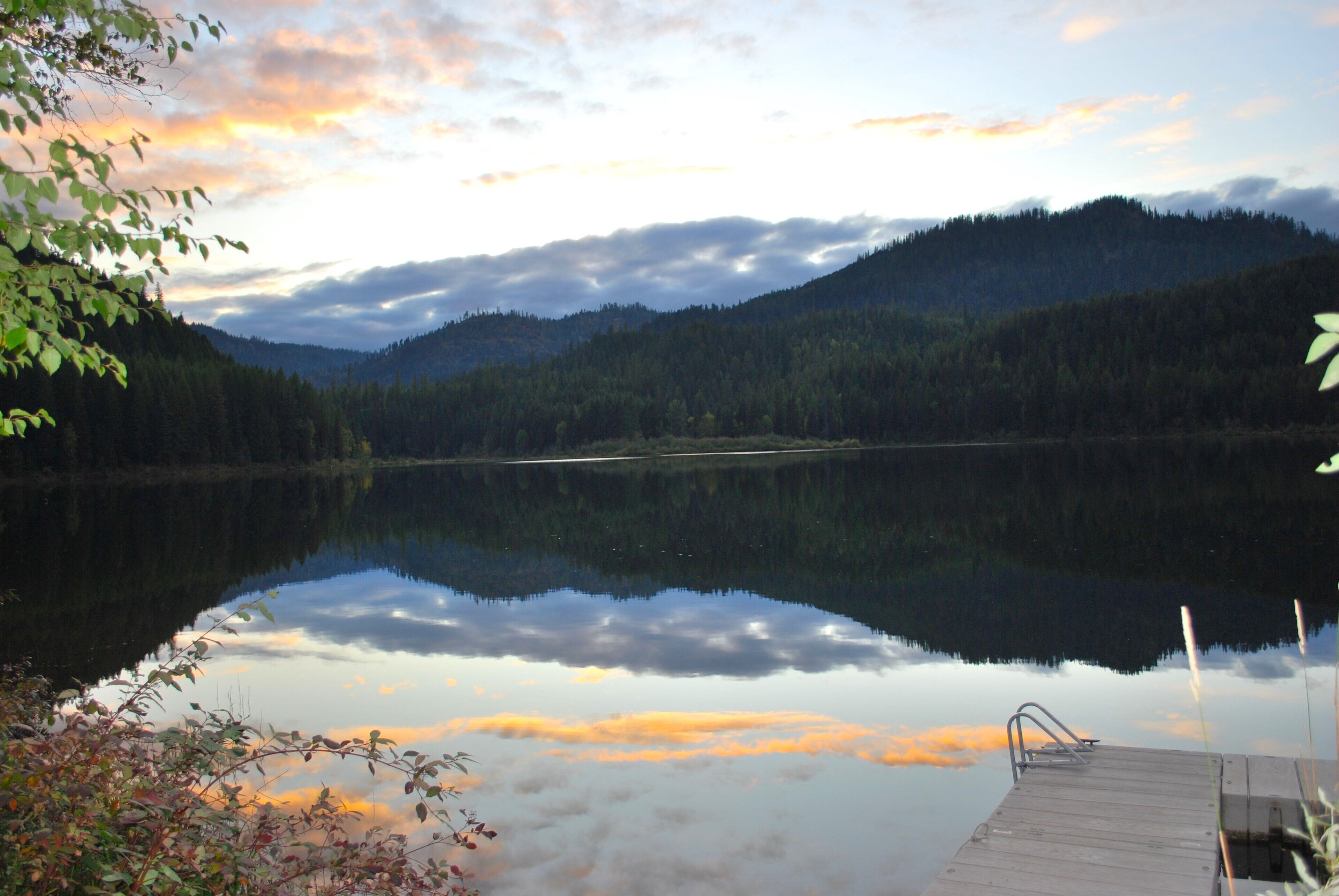 A calm lake with a wooden dock extending into the water, surrounded by dense forest and hills in the background.