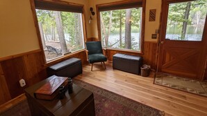 A cozy room with large windows, a green chair, a bench, and a small table with a book and binoculars.