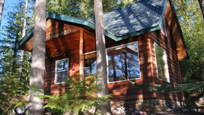 A wooden cabin with a sloped roof, large windows, and a stone foundation surrounded by trees.