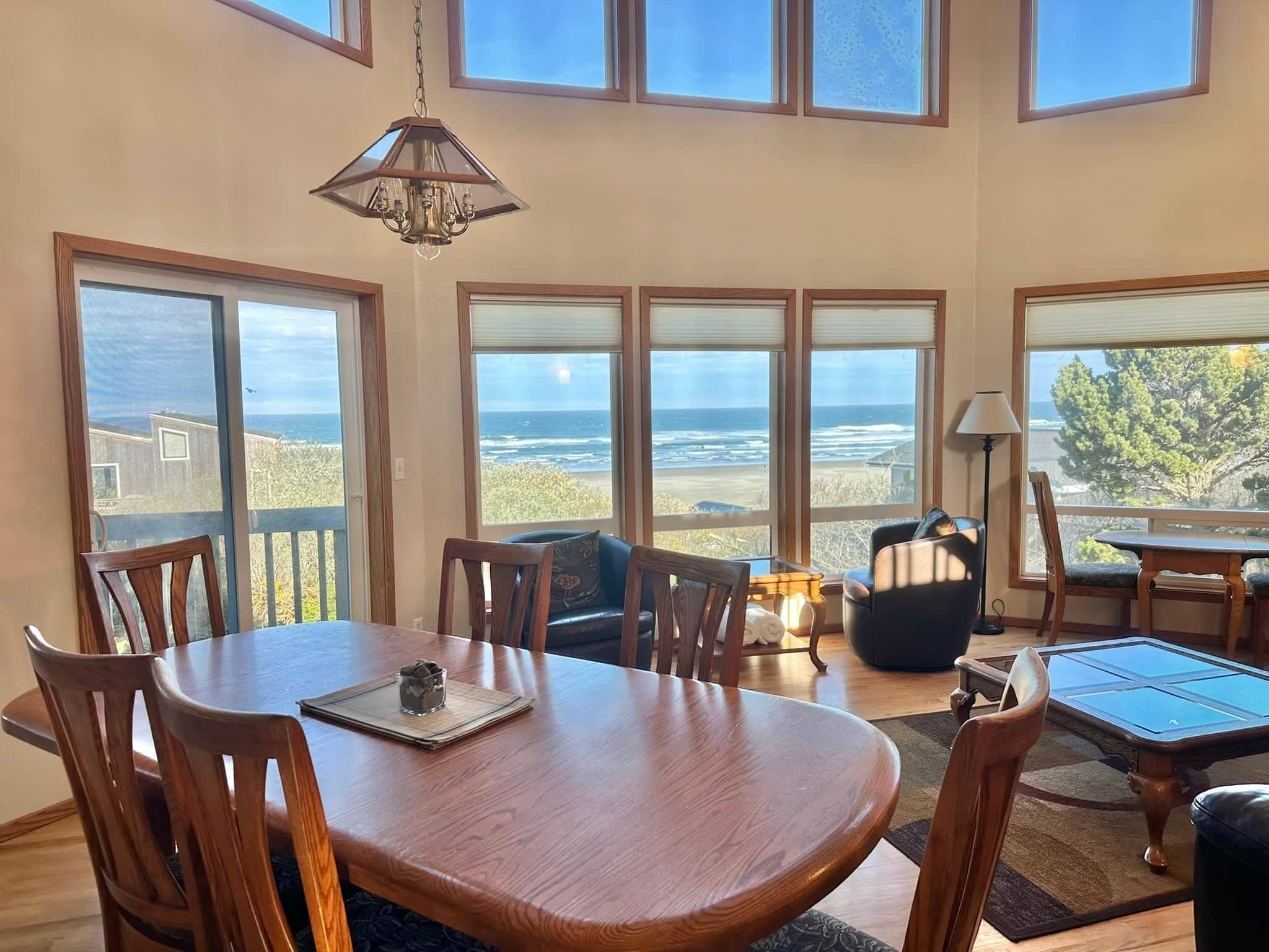 A dining area with a wooden table and chairs, large windows with ocean views, and a hanging light fixture.