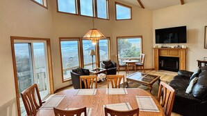 A spacious living room with a large wooden dining table, multiple chairs, a fireplace, a television, and large windows offering ocean views. - Pacific Panorama - 5 bedrooms + loft (Bandon)