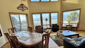 A spacious living room with a large wooden dining table, comfortable seating, and large windows offering ocean views. - Pacific Panorama - 5 bedrooms + loft (Bandon)
