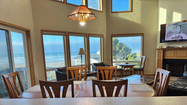 A dining area with a view of the ocean, wooden furniture, and a television on the wall. - Pacific Panorama - 5 bedrooms + loft (Bandon)